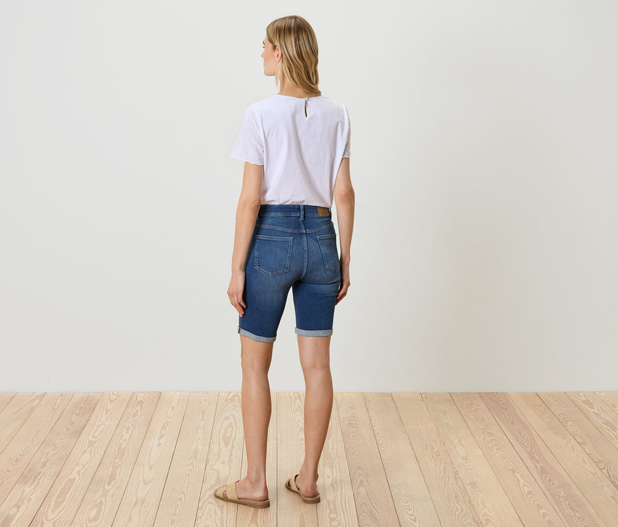 Une femme se tient dos à la caméra, portant un t-shirt blanc, un short en jean et des sandales.
