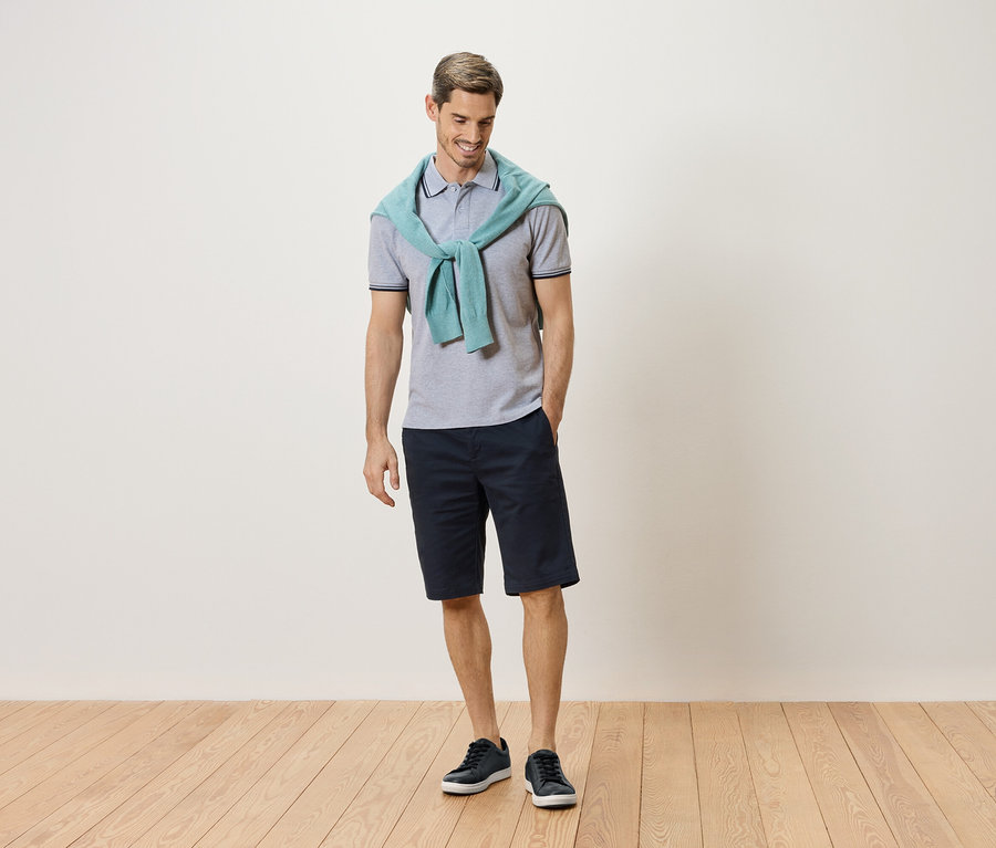 Un homme debout avec un pull bleu clair autour du cou, un polo gris, un short bleu marine et des baskets bleues.