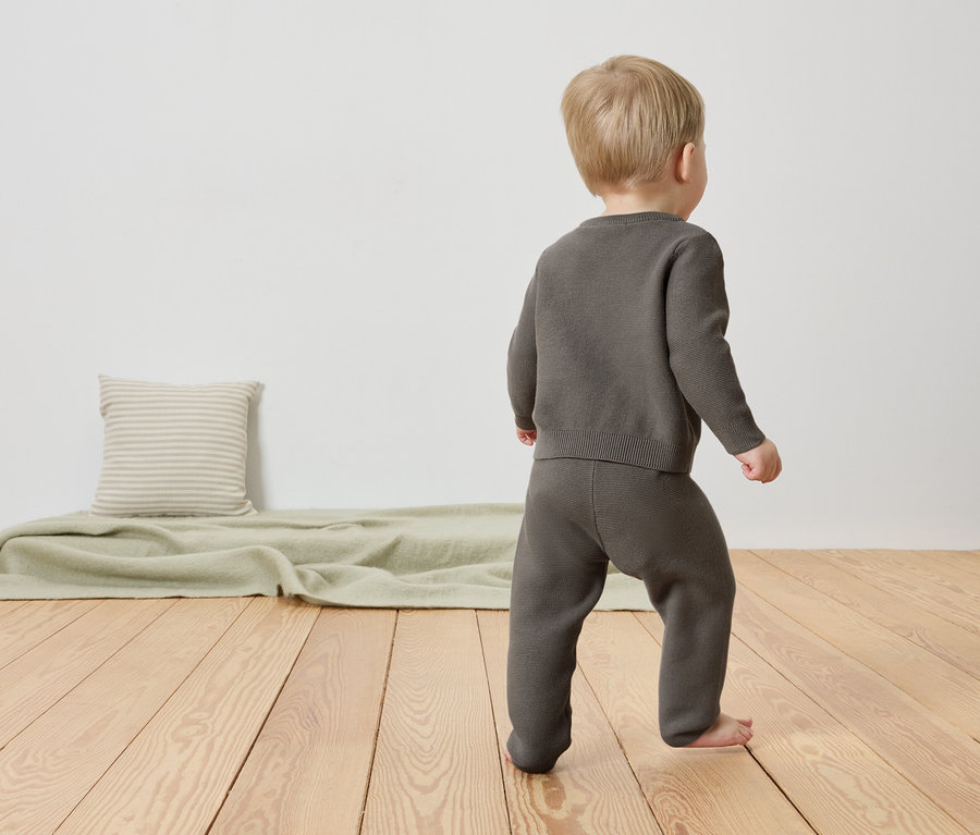 Un jeune enfant marche pieds nus sur un plancher en bois. Il porte un ensemble en tricot gris composé d'un pull et d'un pantalon.