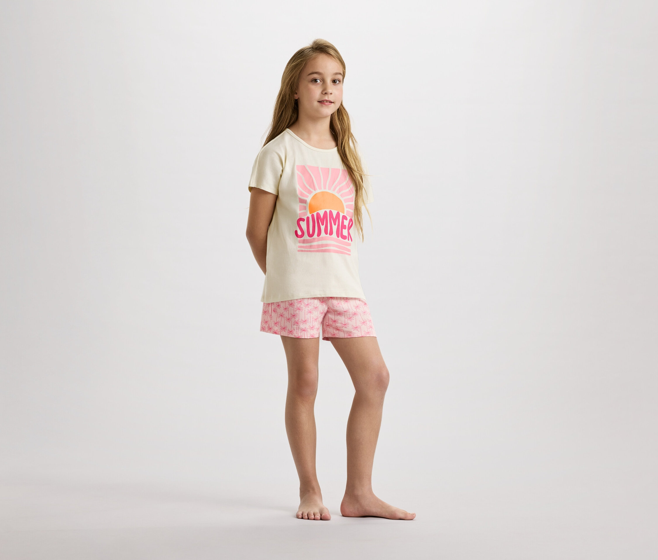 Une jeune fille se tient pieds nus sur un fond blanc, portant un t-shirt beige avec un imprimé rose "Summer" et un short à motifs de palmiers roses.