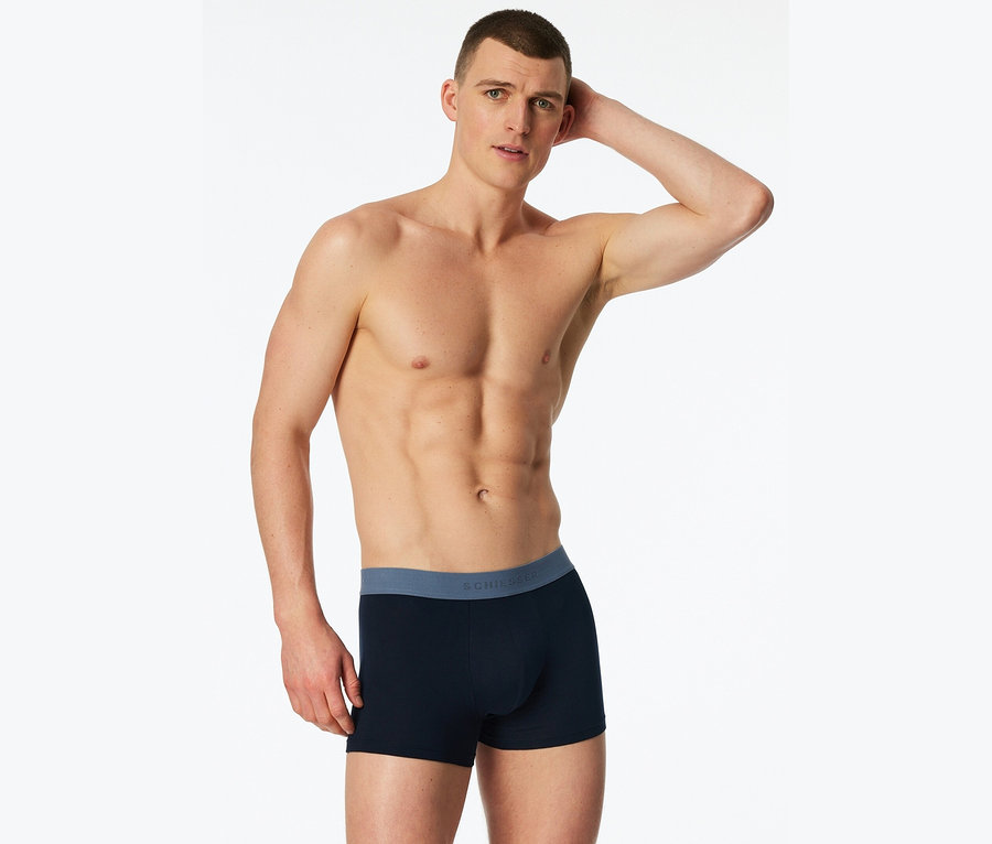 Un homme torse nu pose en caleçon boxer bleu marine avec une ceinture bleue sur fond blanc.