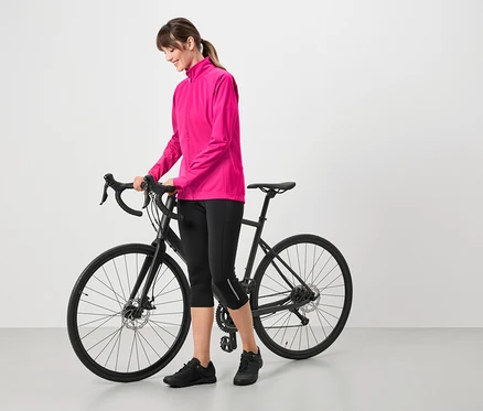 Femme portant une veste rose, un corsaire de cycliste noir et des chaussures noires, debout à côté d'un vélo noir.