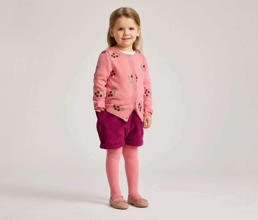 Une fillette blonde porte un cardigan rose à motifs de cerises, un short violet, des collants roses et des ballerines.