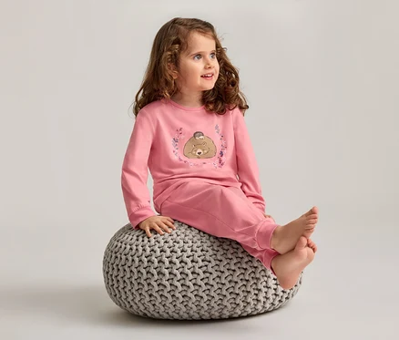 Une fillette aux cheveux bouclés est assise pieds nus sur un pouf tricoté gris et porte un pyjama rose avec un motif d'ours.