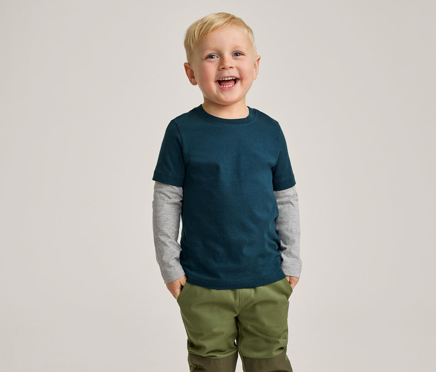 Un jeune garçon blond souriant porte un t-shirt bleu sarcelle foncé sur un t-shirt gris à manches longues et un pantalon vert olive, les mains dans les poches.