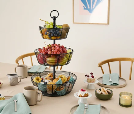 Un panier à fruits à trois étages en fil métallique est posé sur une table avec des pâtisseries, des fruits et de la vaisselle.
