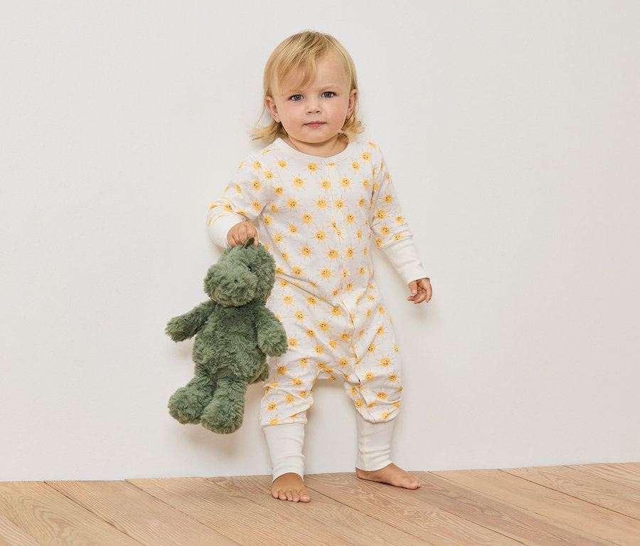 Un enfant blond en combinaison à motifs de soleil tient un dinosaure en peluche vert.