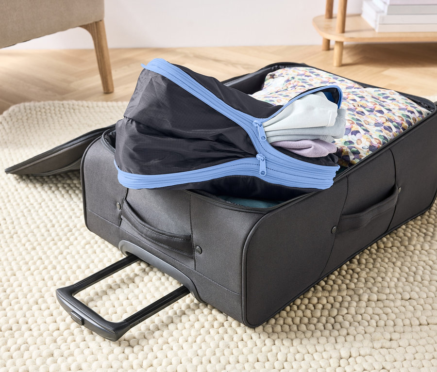 Valise grise ouverte sur un tapis, remplie de vêtements et d'un cube d'emballage noir avec fermeture éclair bleue.