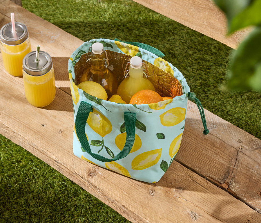 Deux boissons dans des verres et un sac isotherme à motif citron rempli de bouteilles et d'agrumes sont posés sur un banc en bois devant une pelouse verte.