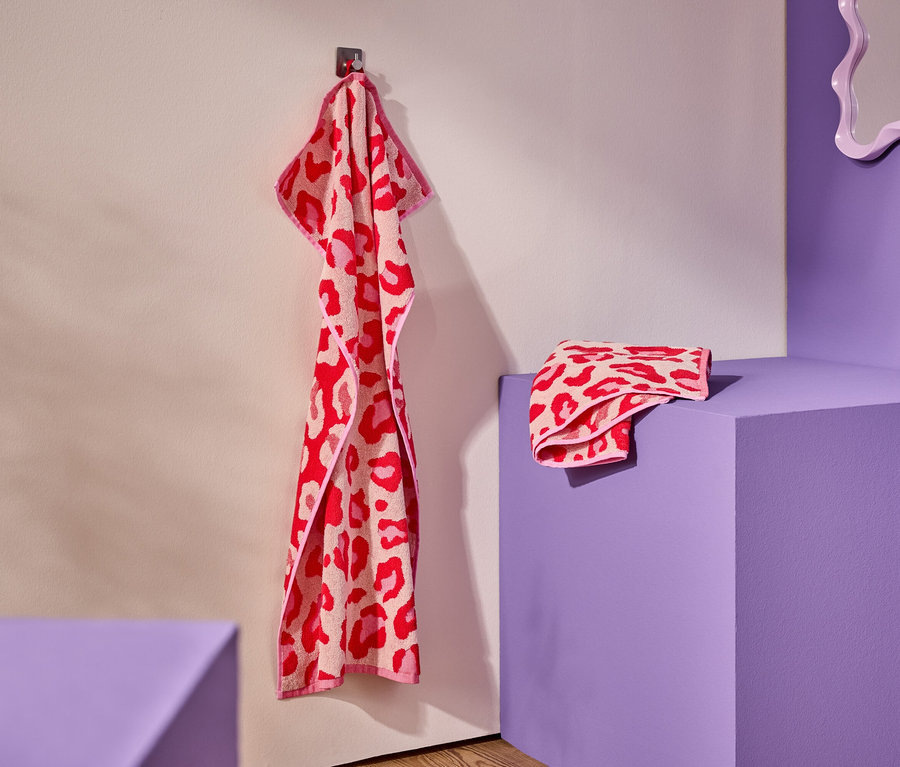 Une serviette rose et rouge à motif léopard est suspendue à un crochet. Une serviette pliée repose sur un piédestal violet.