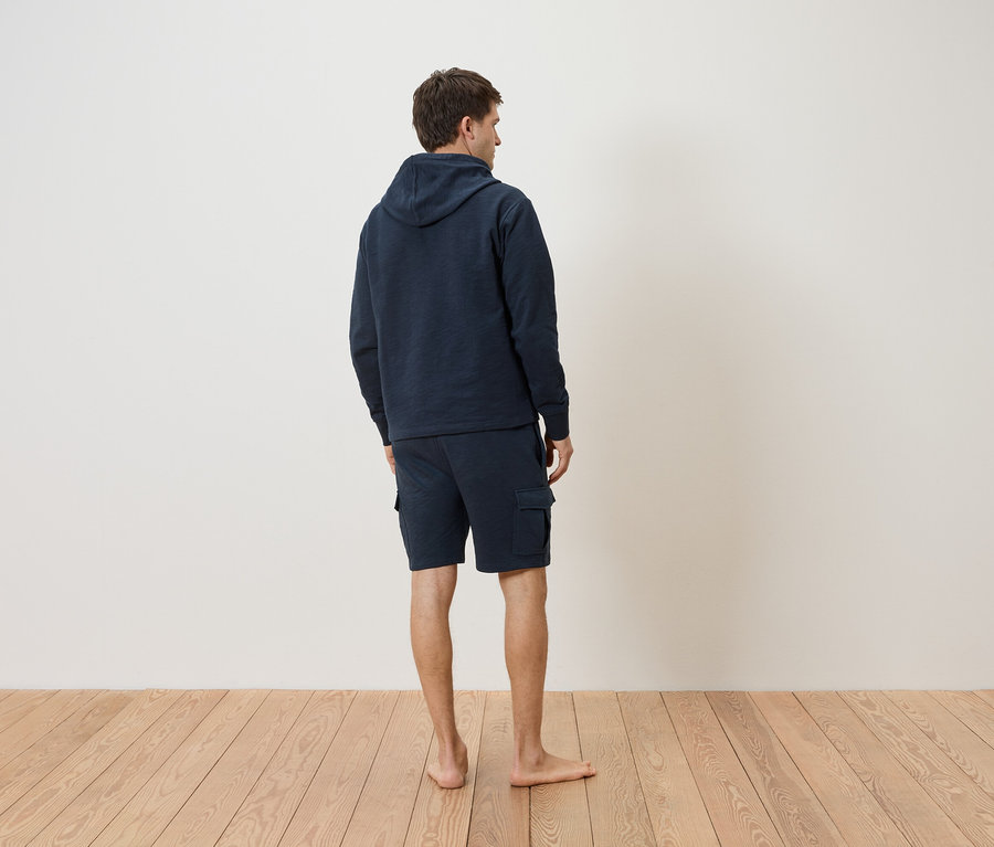 Homme vu de dos, portant un sweat à capuche bleu marine et un short sur un plancher en bois.