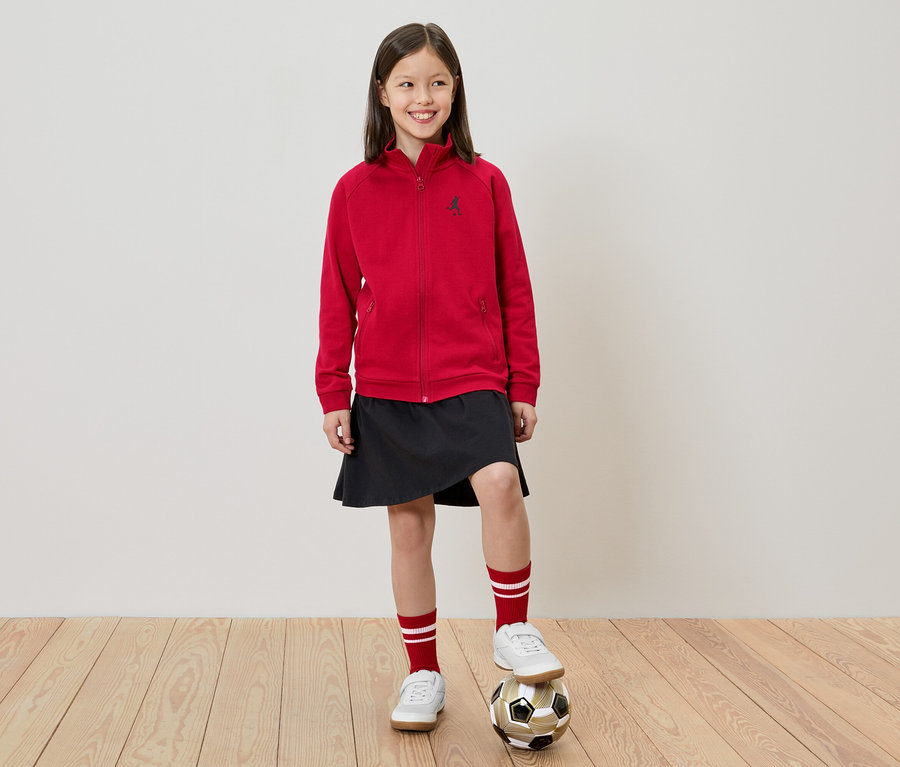 Une fille se tient un pied sur un ballon de foot, porte une veste rouge, une jupe noire, des chaussures blanches et des chaussettes rouge et blanche.