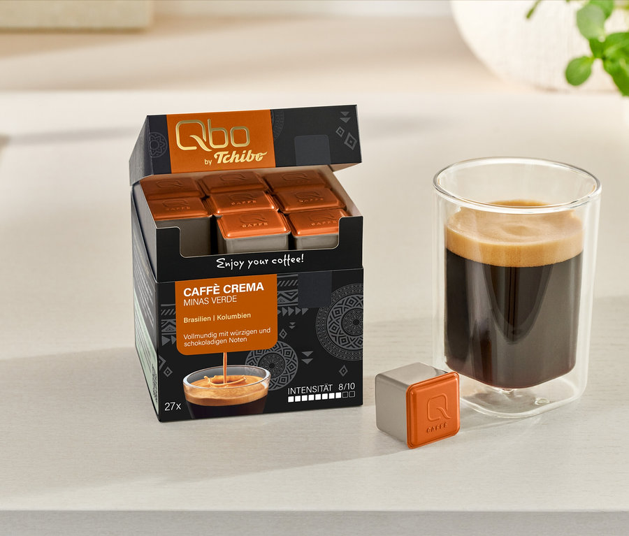 Une boîte ouverte de capsules Qbo Caffè MINAS VERDE, une capsule individuelle et un verre de café sont posés sur un comptoir.