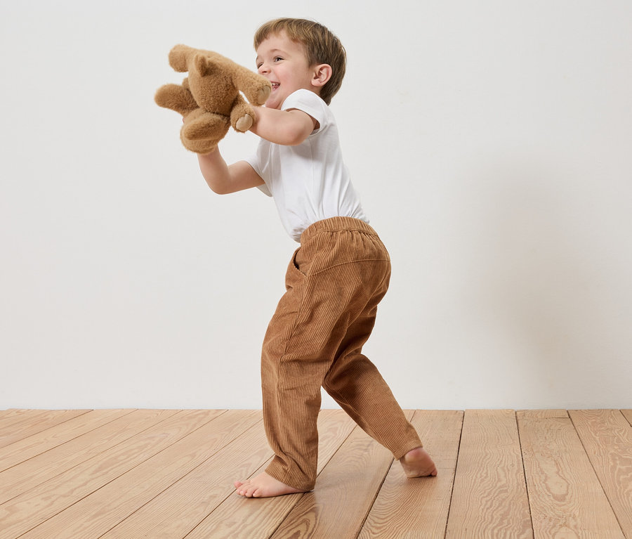 Un garçon en pantalon de velours côtelé et t-shirt joue pieds nus avec un ours en peluche devant un mur blanc.