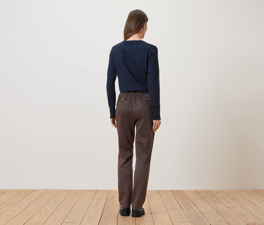 Une femme vue de dos, portant un haut bleu marine et un pantalon à motifs, se tient sur un plancher en bois devant un mur blanc.
