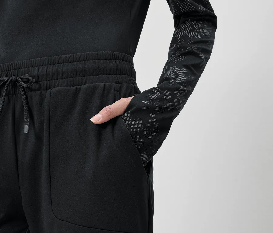 Femme avec une main dans la poche d'un pantalon de jogging noir à cordon et un haut noir à manches longues à motifs.