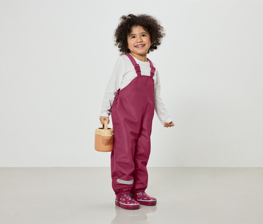 Un enfant souriant aux cheveux bouclés porte une salopette bordeaux et des bottes en caoutchouc à motifs floraux, et tient un arrosoir marron clair.