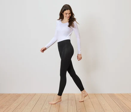 Une femme pieds nus, portant un haut blanc à manches longues et un legging noir, marche sur un plancher en bois devant un mur blanc.