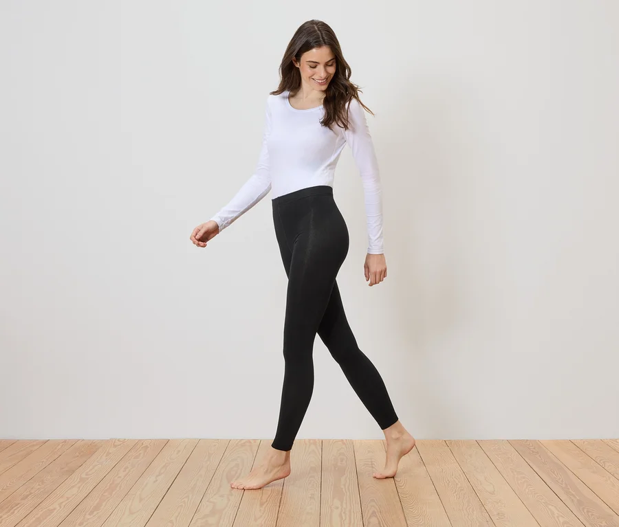 Une femme pieds nus, portant un haut blanc à manches longues et un legging noir, marche sur un plancher en bois devant un mur blanc.