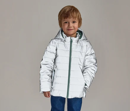 Un garçon souriant aux cheveux blonds porte un blouson argenté réfléchissant à capuche avec une fermeture éclair verte sur un jean bleu.