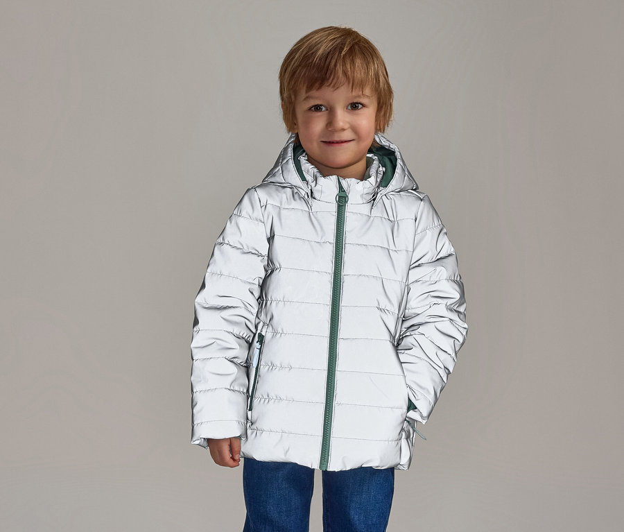 Un garçon souriant aux cheveux blonds porte un blouson argenté réfléchissant à capuche avec une fermeture éclair verte sur un jean bleu.