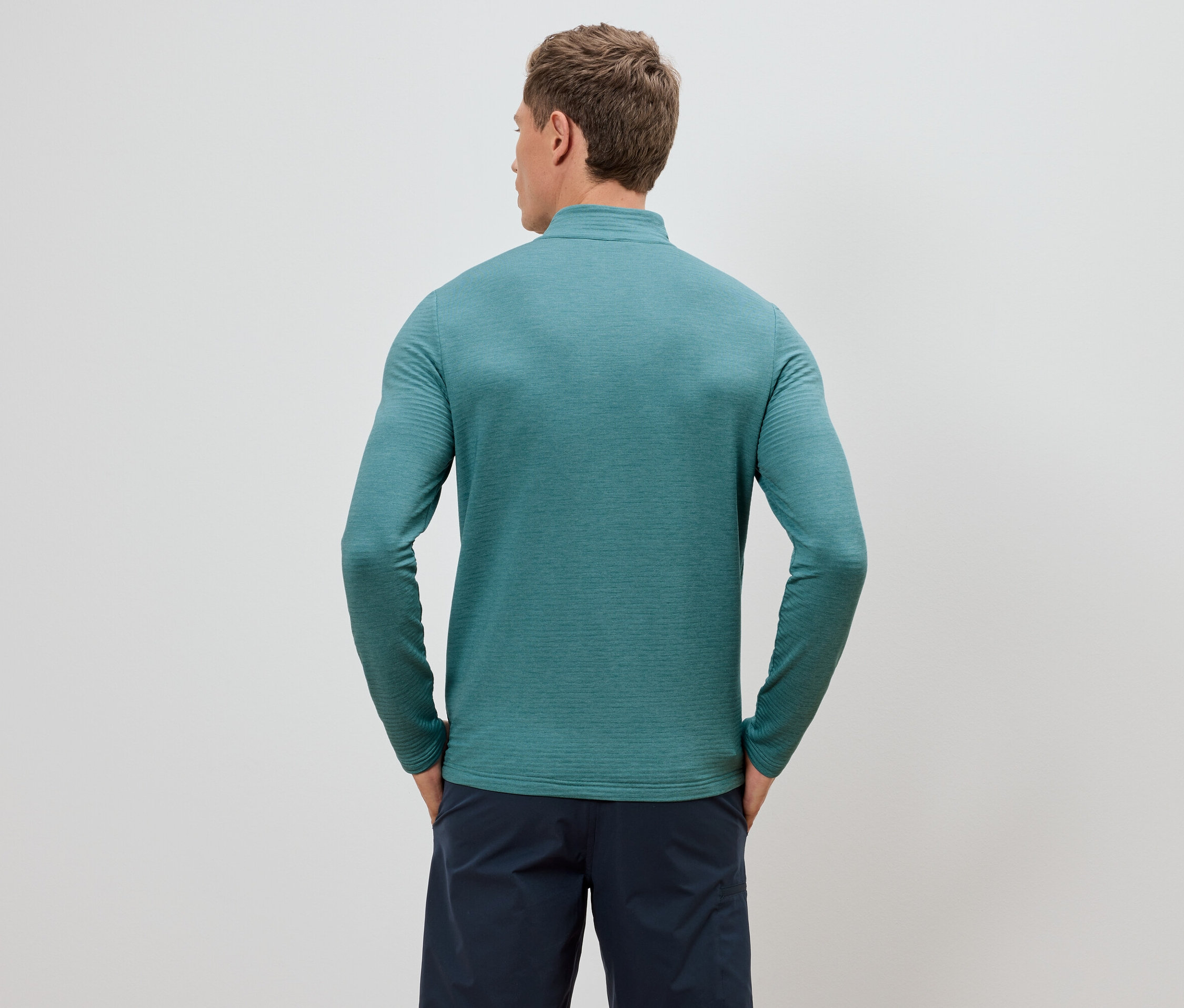 Homme vu de dos portant un haut turquoise à manches longues et un short bleu marine, les mains dans les poches.