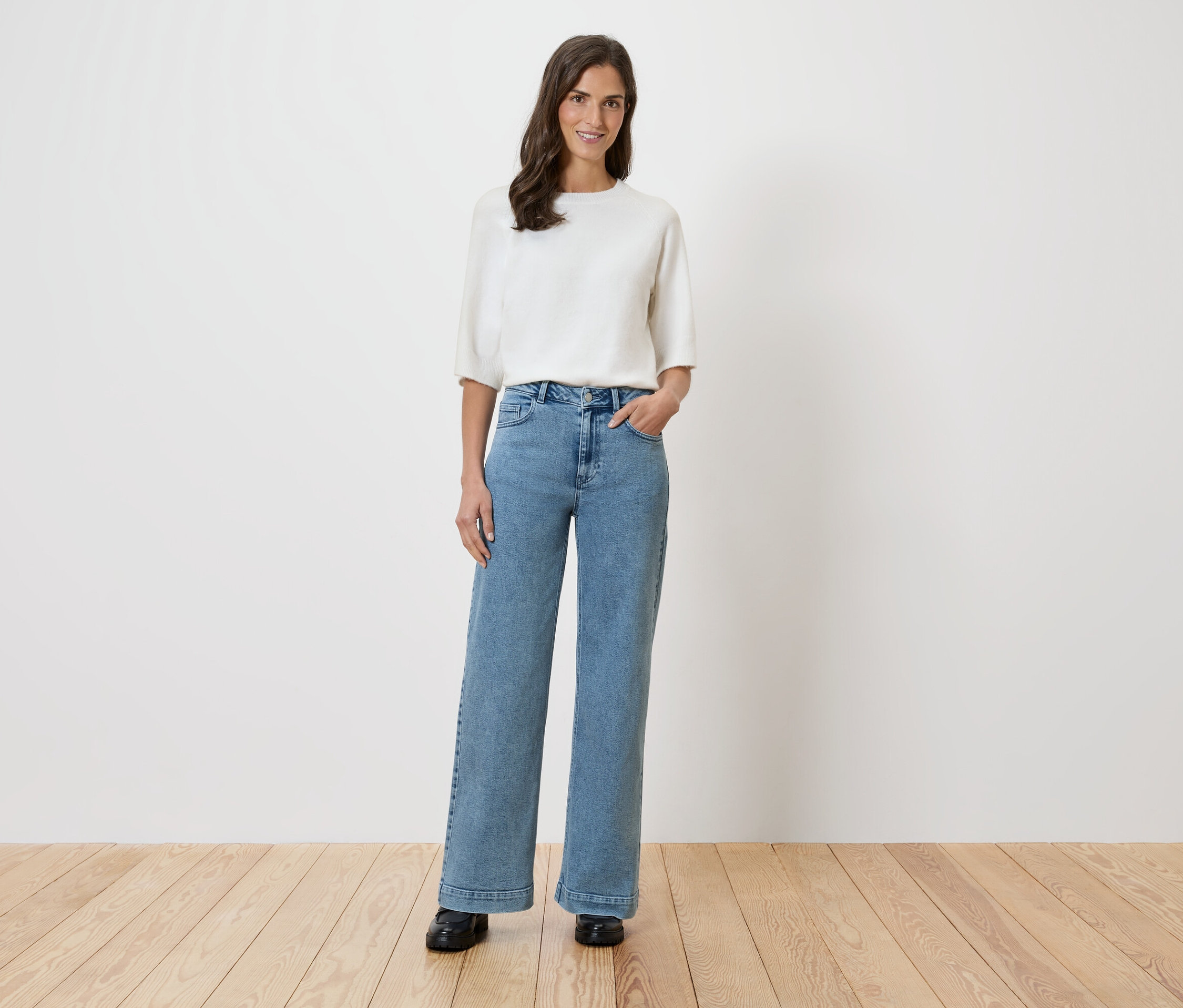 Une femme se tient devant un mur blanc, vêtue d'un chemisier blanc et d'un jean large.