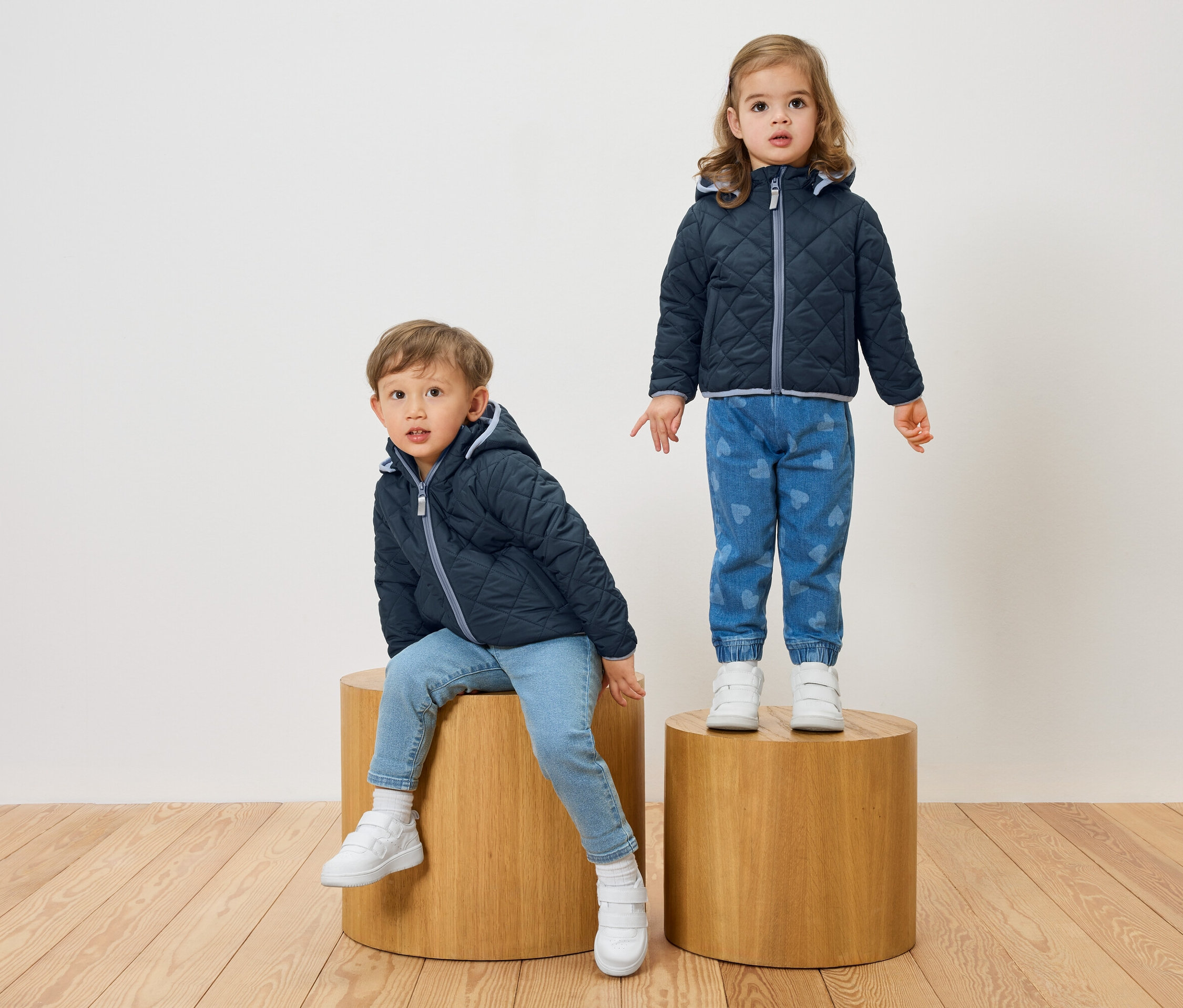 Un garçon est assis sur un tabouret en bois tandis qu'une fille se tient debout sur un autre. Ils portent tous les deux des vestes matelassées bleu marine et des jeans.