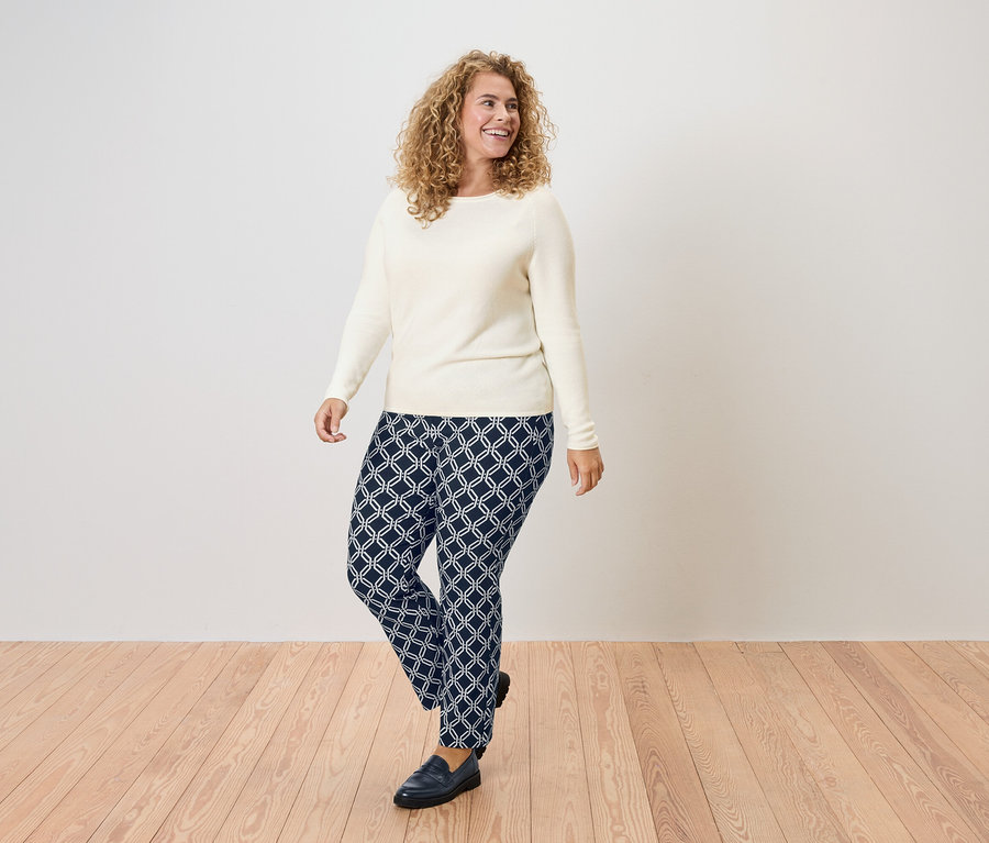 Femme aux cheveux bouclés portant un pull crème et un pantalon bleu marine à motifs géométriques.