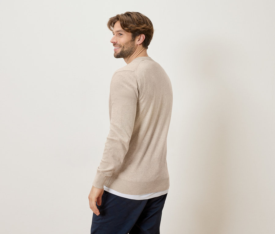 Un homme regarde par-dessus son épaule, portant un pull beige et un pantalon bleu marine sur un fond crème.