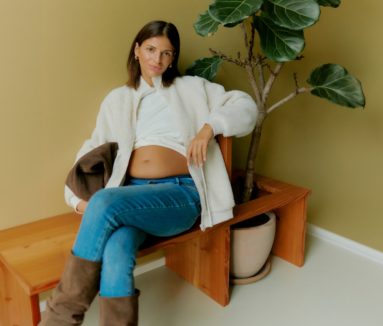 Femme enceinte assise sur un banc à côté d'une plante, portant une veste Teddy 3 en 1 MAMALICIOUS «
MLCARLY » ouverte sur un t-shirt blanc et un jean.