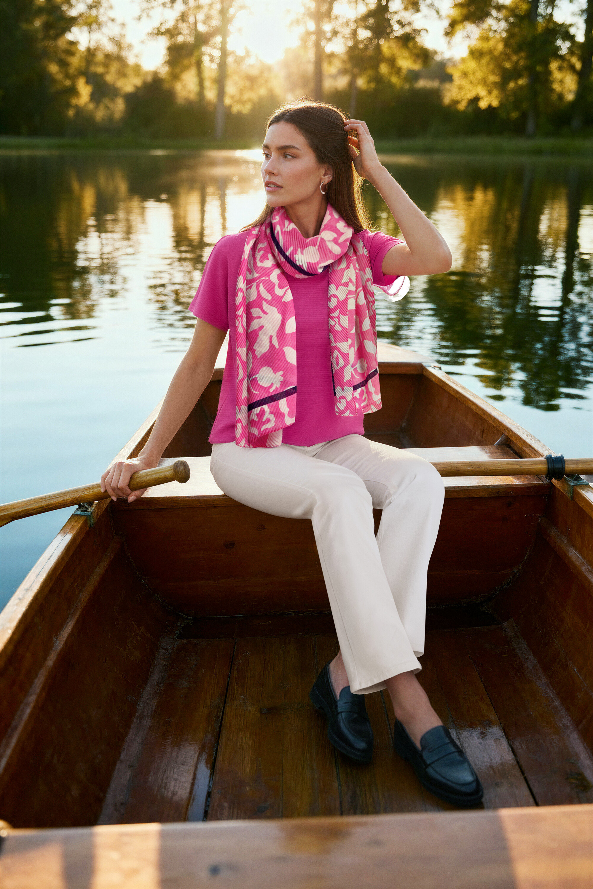 Femme assise dans un bateau, portant un chemisier rose, un jean bootcut – Coupe « Lou », blanc crème, des loafers en cuir et des Créoles vague.