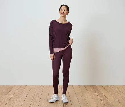 Une femme debout portant un haut à manches longues bordeaux et un legging bordeaux.