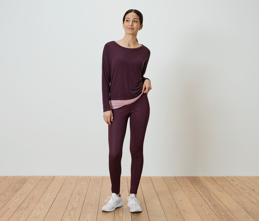 Une femme debout portant un haut à manches longues bordeaux et un legging bordeaux.