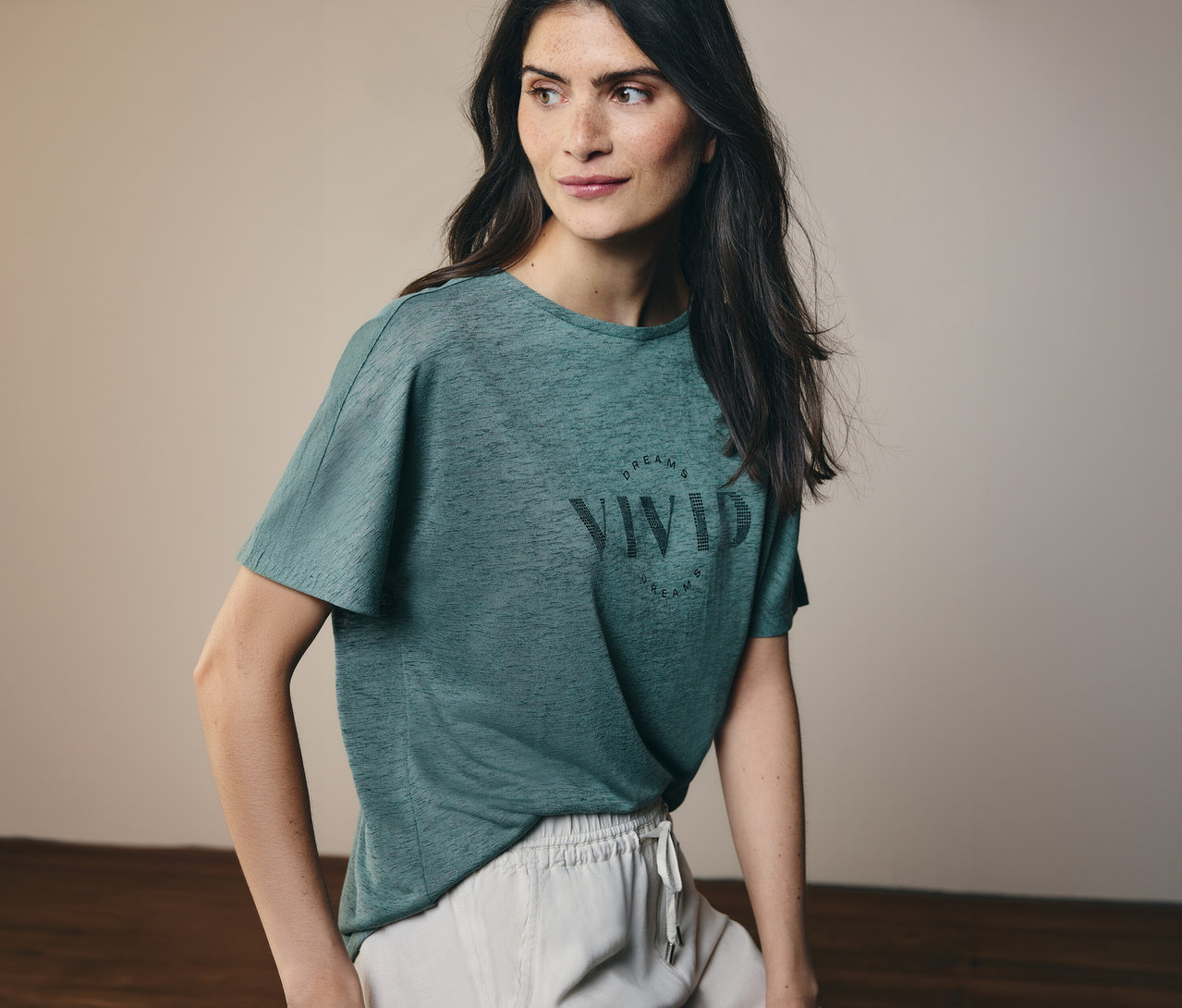 Une femme aux longs cheveux foncés porte un t-shirt vert avec l'inscription "Vivid Dreams" et un short clair.