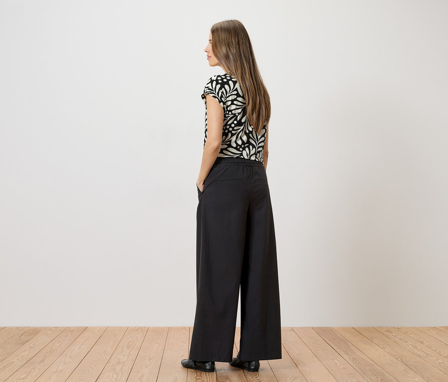 Femme portant un haut à motifs et un pantalon noir ample, les mains dans les poches devant un mur blanc.