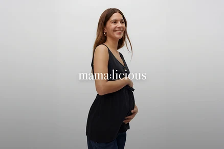 Femme enceinte souriante aux longs cheveux bruns portant un haut de grossesse noir sans manches boutonné et un jean.
