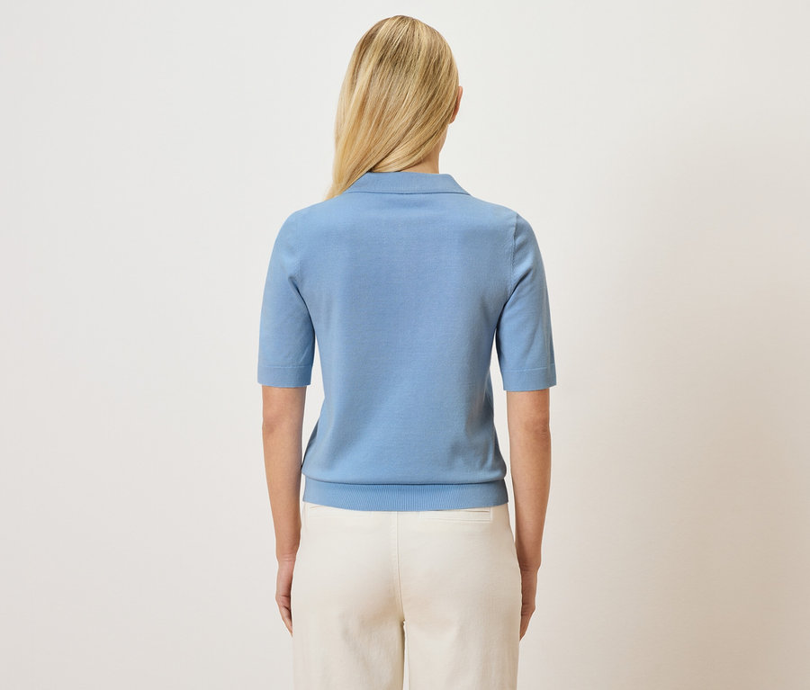 Vue arrière d'une femme blonde portant un polo bleu clair et un pantalon clair.