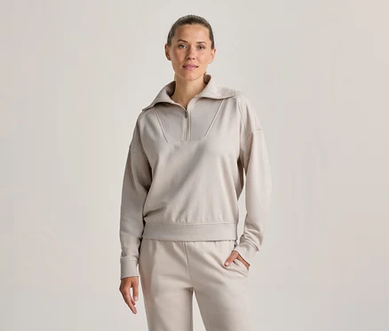 Femme debout, portant un sweatshirt beige clair à demi-zip et un pantalon de jogging assorti, une main dans la poche, regardant devant elle.