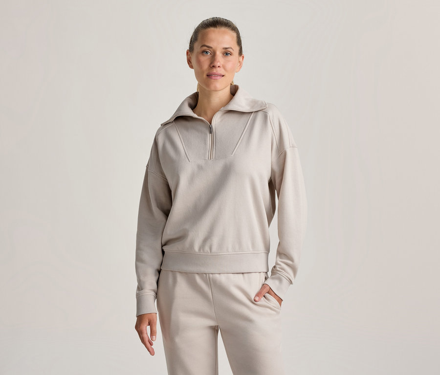 Femme debout, portant un sweatshirt beige clair à demi-zip et un pantalon de jogging assorti, une main dans la poche, regardant devant elle.