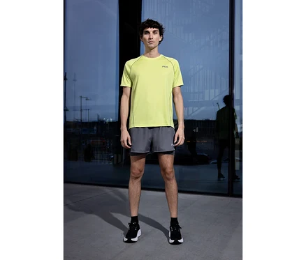 Un homme se tient devant une façade vitrée, portant un T-shirt technique pour homme FILA vert clair, un short gris et des chaussures de sport noires.