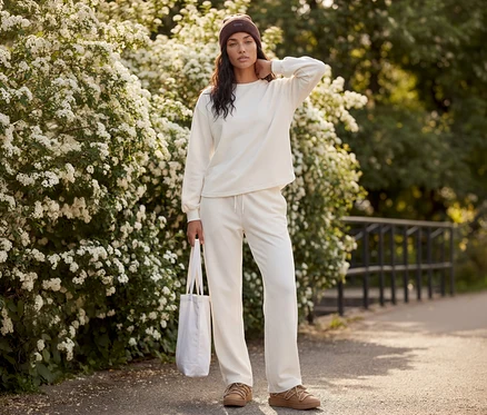 Une femme en survêtement crème, bonnet marron et chaussures marron, tenant un sac en tissu blanc, devant des buissons fleuris.