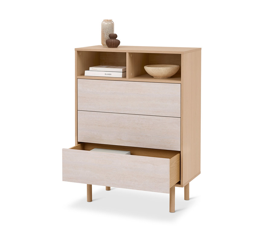 Commode en bois clair avec deux étagères ouvertes et trois tiroirs. L'étagère supérieure contient deux livres et un bol clair, tandis que le tiroir inférieur est légèrement ouvert, révélant un objet blanc. Deux vases décoratifs sont posés dessus.