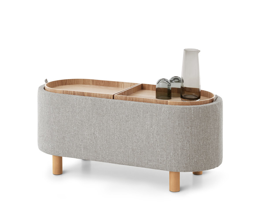Ottoman ovale en tissu gris clair avec quatre pieds en bois, doté de deux plateaux en bois amovibles sur le dessus. Un plateau contient une carafe en verre gris et deux verres assortis.