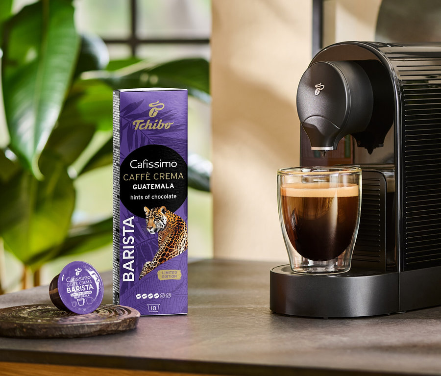 Une machine à café prépare un Cafissimo BARISTA Caffè Crema Guatemala dans un verre. À côté se trouvent un paquet de 10 capsules et une capsule.