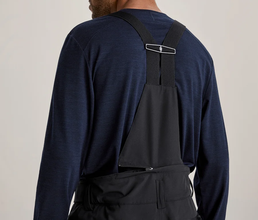 Vue de dos d'une personne portant un t-shirt à manches longues bleu foncé et une salopette noire avec des bretelles se rejoignant par une boucle argentée.