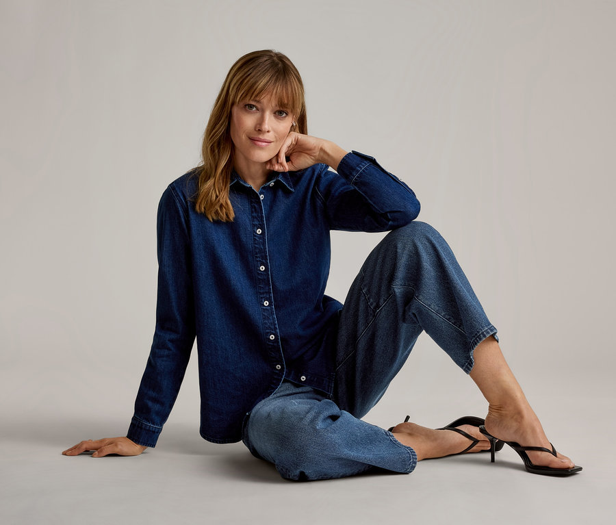 Femme assise sur le sol, portant une chemise en jean bleu foncé, un pantalon en jean court bleu clair et des sandales noires à talons hauts, regardant l'appareil photo, la main sur le menton.