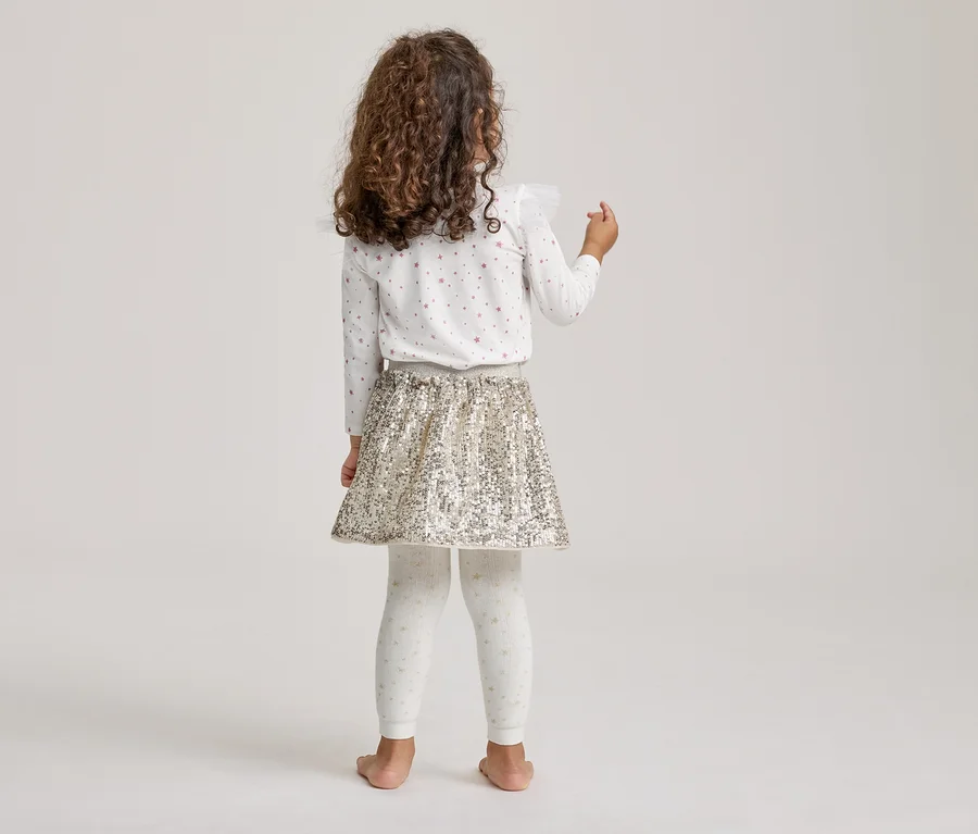 Enfant debout dos à la caméra, portant un haut blanc à manches longues avec des étoiles, une jupe à sequins dorés et un legging blanc à motifs.