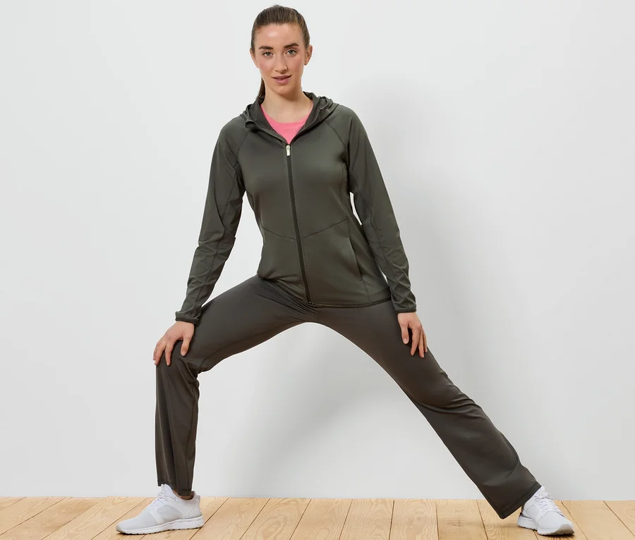 Femme en survêtement vert olive s'étirant sur un plancher en bois devant un mur blanc.