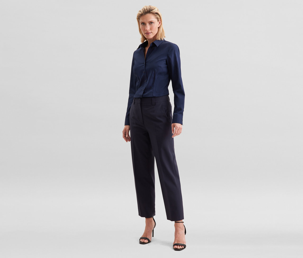 Femme blonde debout, portant une chemise à manches longues bleu marine, un pantalon cropped bleu marine et des sandales noires à talons hauts.